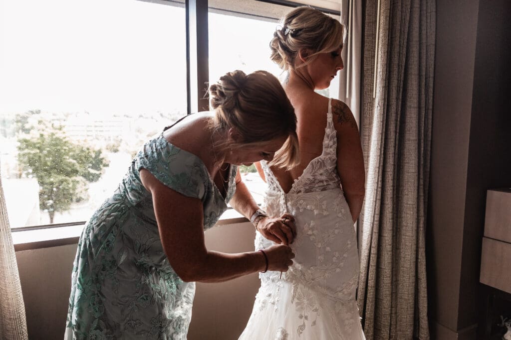 Pittsburgh bride standing by hotel window as her mom helps her into her wedding dress