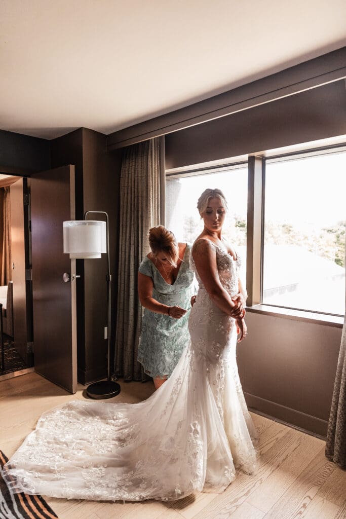 Mother of the bride helping her daughter into her wedding dress by the window at Pittsburgh Airport Marriott