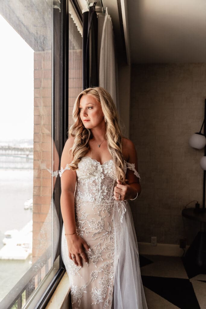 Bride looking out a window framed by hotel curtains before her wedding