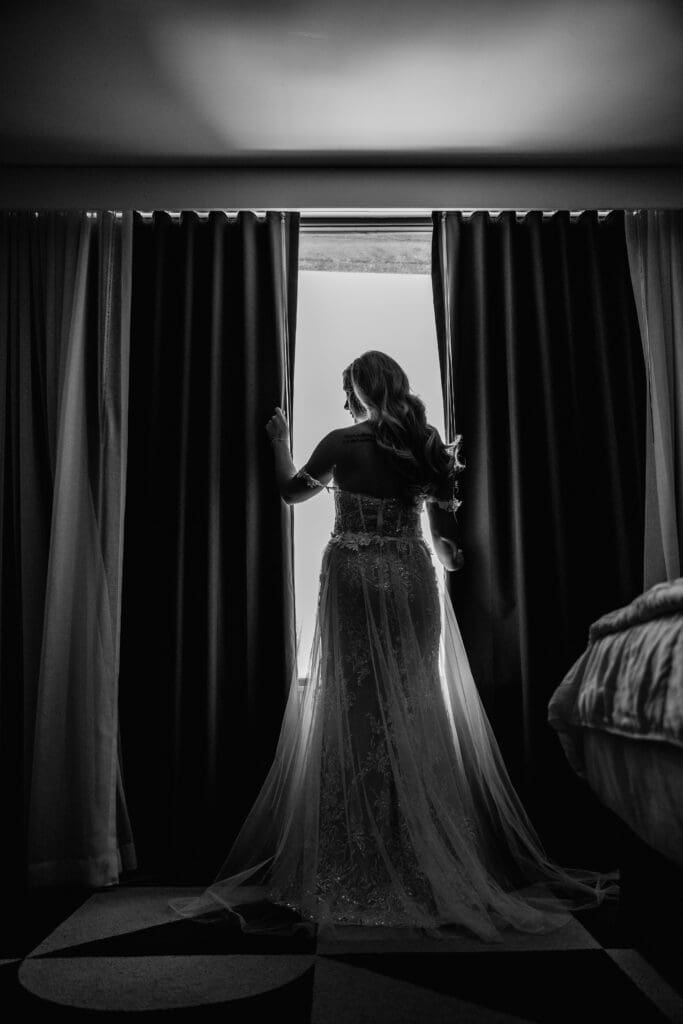 Silhouette of a bride standing in front of a window framed by hotel curtains before her wedding