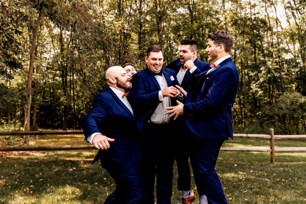 Groomsmen in navy blue suits jumping and laughing during wedding portraits at The Barn at Ever Thine in Butler County, PA