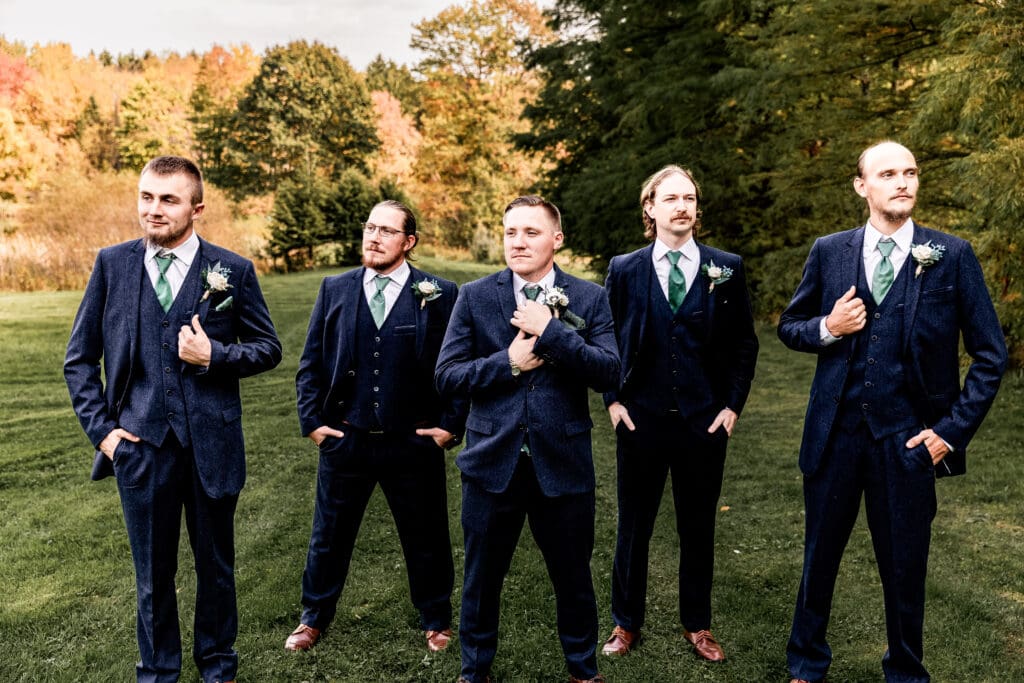 Groom and groomsmen adjusting jackets in front of fall foliage and pond at Pinehall at Eisler Farm
