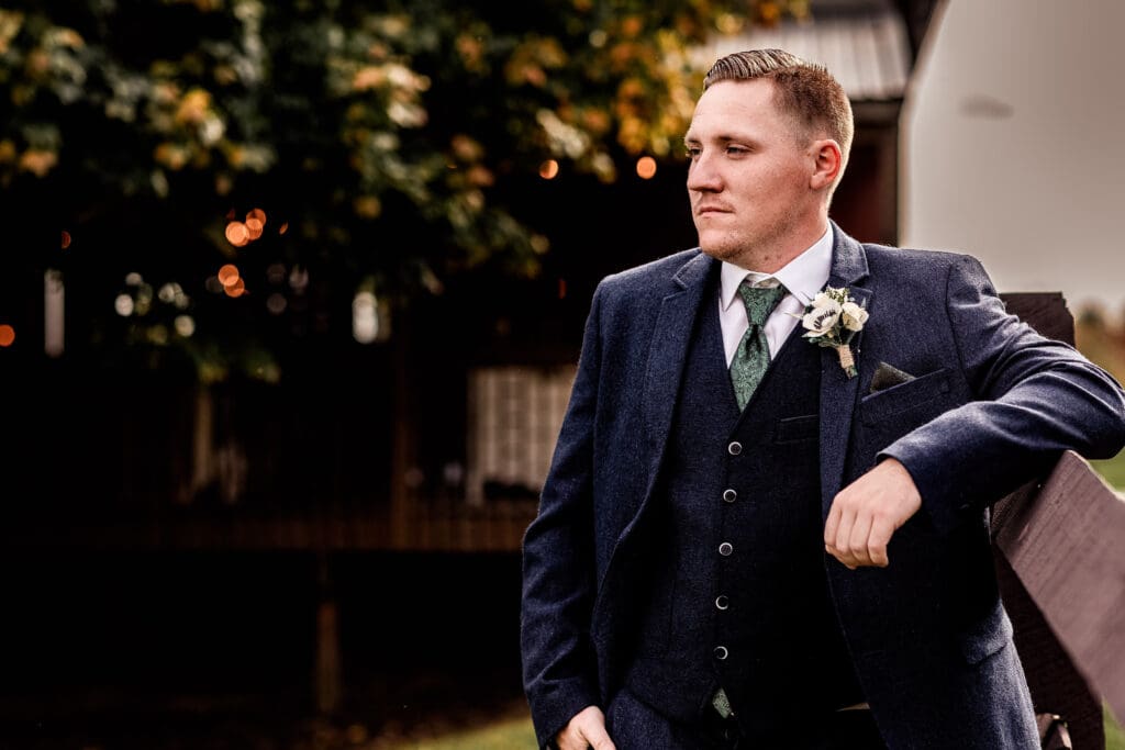 Groom leaning against fence with Pinehall at Eisler Farm reception barn behind him