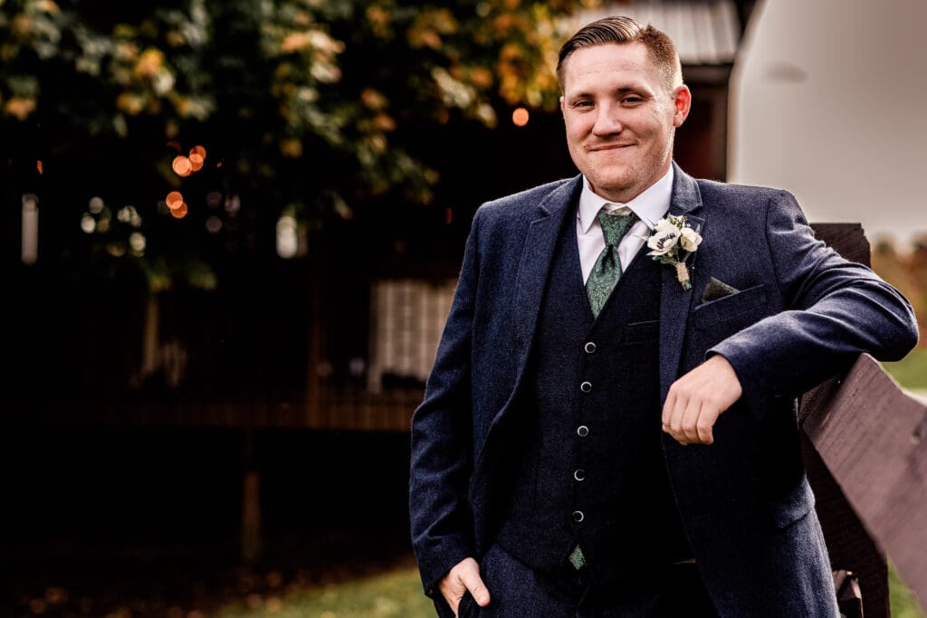 Groom casually leaning on fence with reception barn in background at Pinehall at Eisler Farm