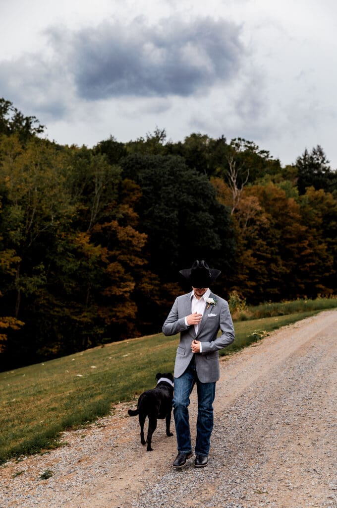 Groom in jeans walking black dog along gravel path with autumn trees at Hinckston Run Farm