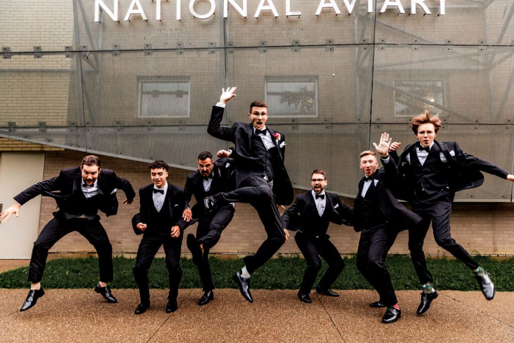 Groomsmen performing jumping pose against outdoor wall at the National Aviary wedding