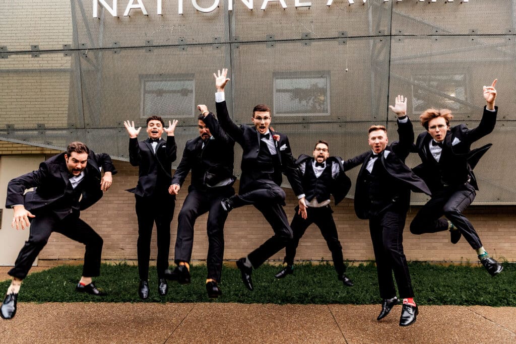 Groomsmen in black suits jumping together during wedding portraits at the National Aviary