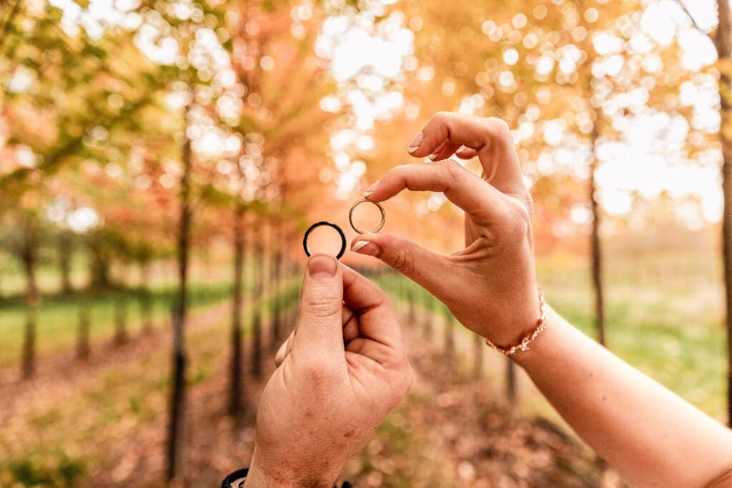 Close-up of bride and groom holding wedding bands with fall foliage in background at Pinehall at Eisler Farm