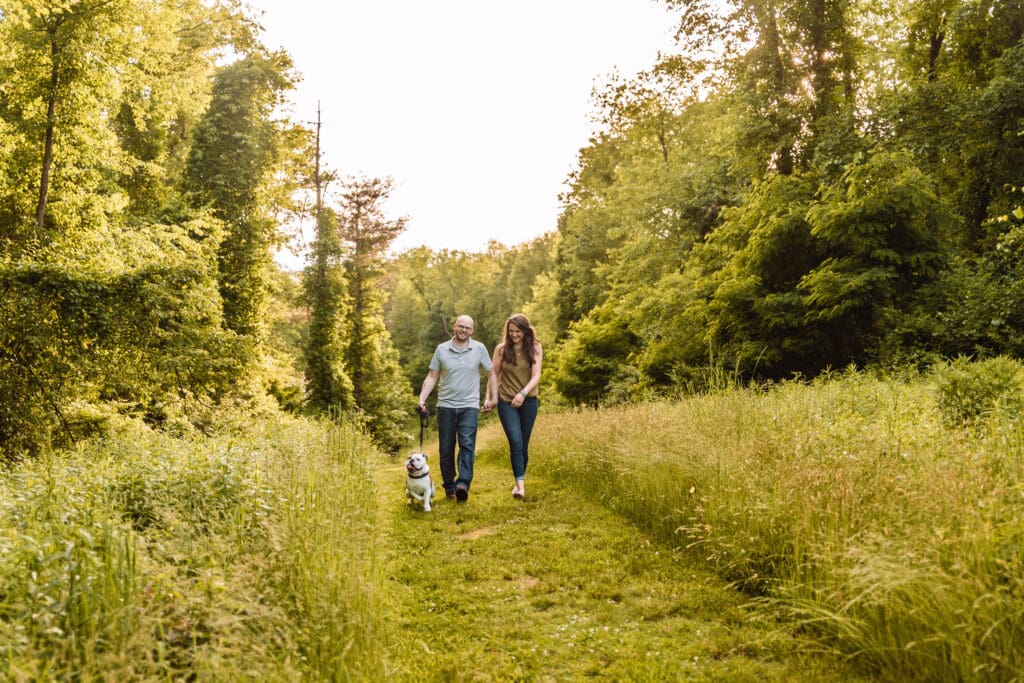 Couple runs together with their dog on a grassy path surrounded by tall autumn grasses during a sunny engagement session