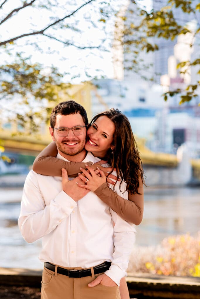 Happy couple shares playful moments outdoors near a river during a Pittsburgh spring engagement photoshoot