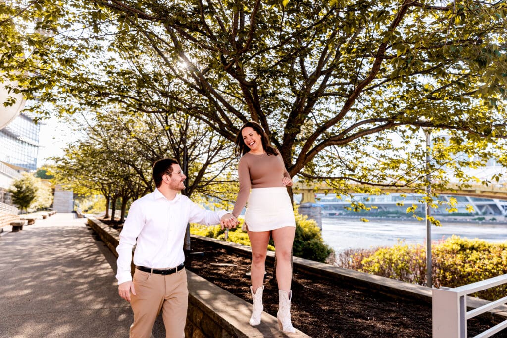 Couple shares a playful moment along a tree-lined waterfront promenade in Pittsburgh on a sunny day