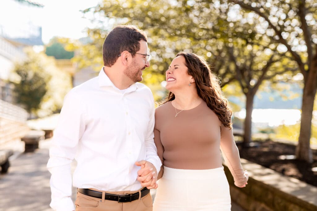 Couple strolls along a scenic Pittsburgh riverside path beneath golden spring trees during an engagement session