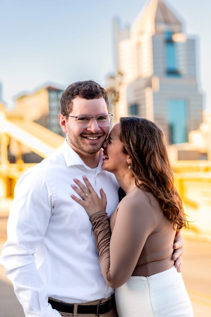 Romantic couple shares tender moments on a bridge with the Pittsburgh city skyline behind them during an engagement session