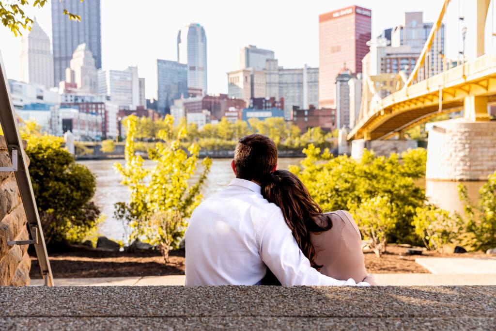 Couple sits together on a stone wall overlooking the Pittsburgh skyline during golden hour engagement photos