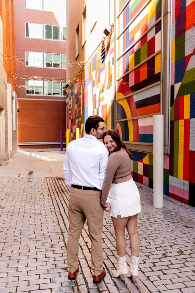 Couple snuggles together in an urban Pittsburgh alleyway featuring a colorful geometric mural, brick paving stones, and white columns