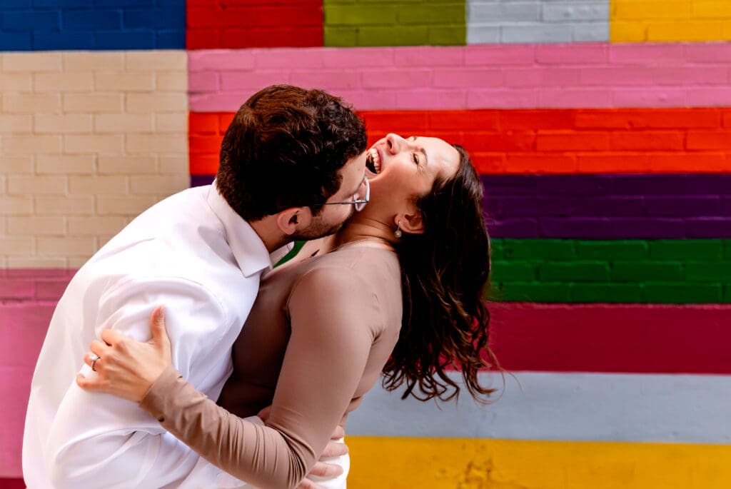 Two people share a passionate embrace against a vibrant colorful striped wall during an engagement photo session