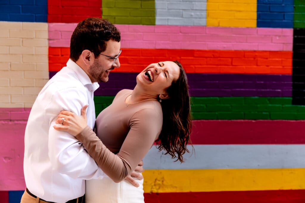 Couple laughs together against a vibrant multicolored striped wall during a fun engagement photoshoot