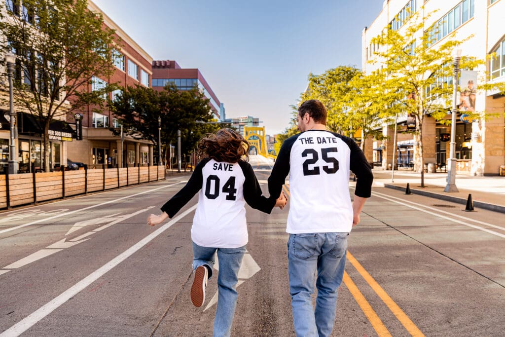 Couple wearing matching raglan shirts and jeans walks side by side on a Pittsburgh sidewalk surrounded by colorful fall foliage