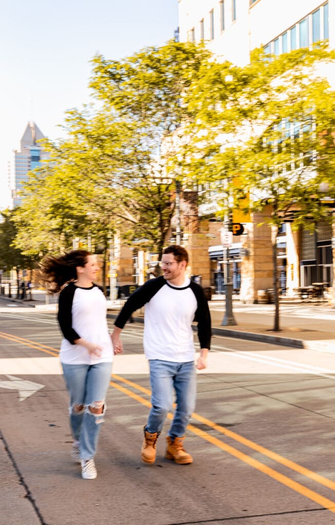 Two people in matching raglan shirts and jeans walk together on a Pittsburgh city sidewalk lined with spring trees during an engagement session