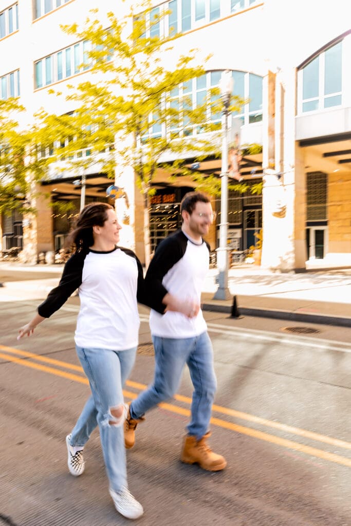 Two people walk together in matching raglan shirts and jeans along a tree-lined Pittsburgh city sidewalk during an spring engagement photoshoot