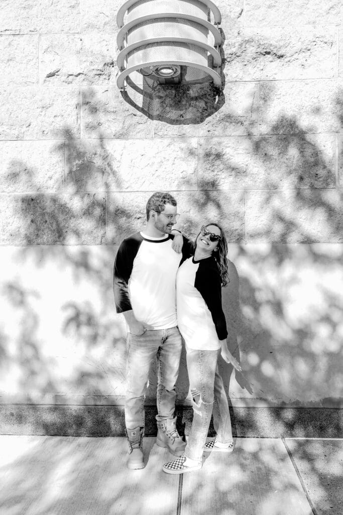 Couple in matching baseball-style shirts leans against a beige wall with dappled shadows and an industrial light fixture during a Pittsburgh engagement shoot