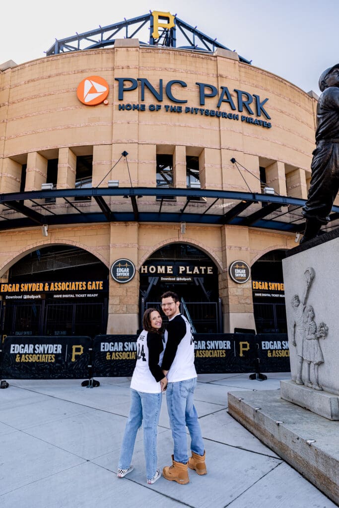 Couple stands in front of the PNC Park baseball stadium entrance with black and gold signage during a Pittsburgh engagement session