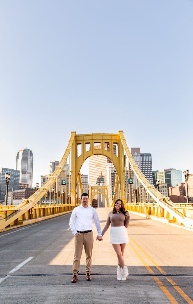 Couple stands on Pittsburgh’s iconic yellow Roberto Clemente Bridge at sunrise during a city engagement session