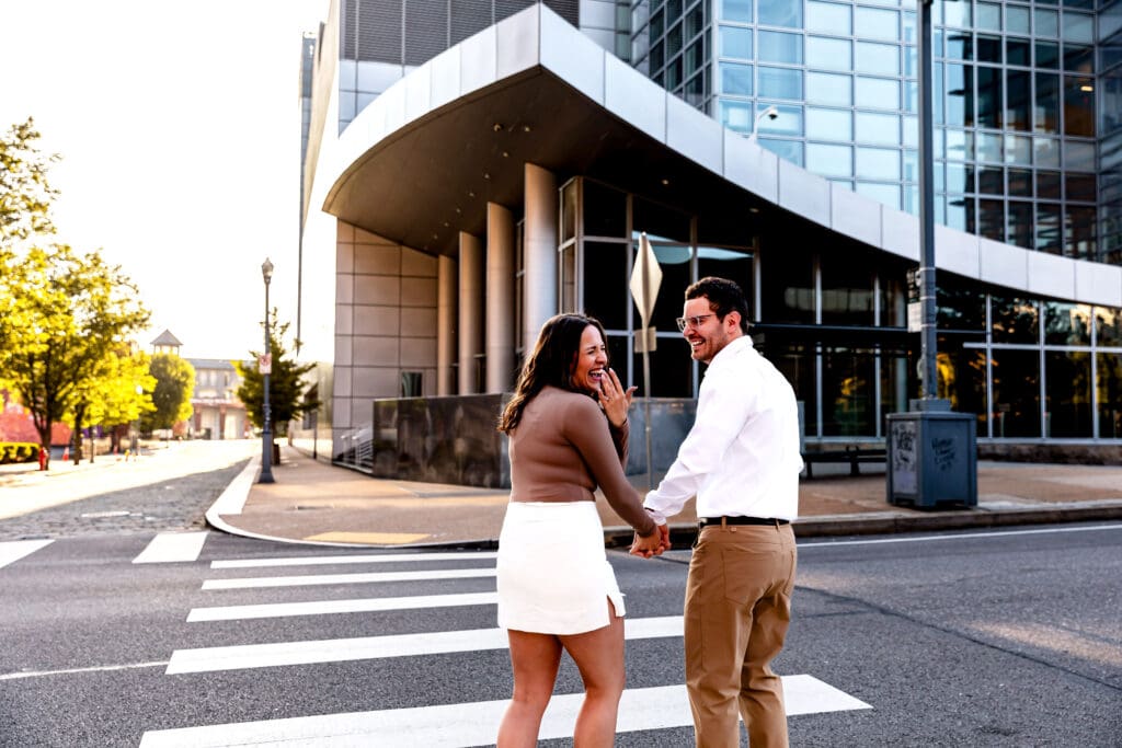 Couple in formal attire stands outside a modern glass office building in Pittsburgh during a sunny engagement photo session