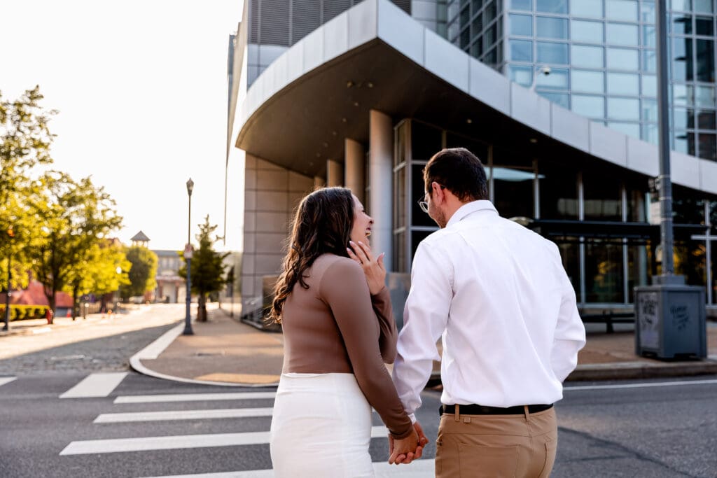 Couple laughs in front of a modern glass office building in Pittsburgh during a romantic engagement photoshoot