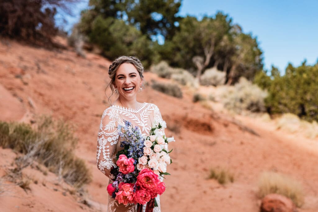 Bride holding a vibrant bouquet and laughing at the camera during her Look Glass Arch Moab elopement, with red rock desert scenery in the background.