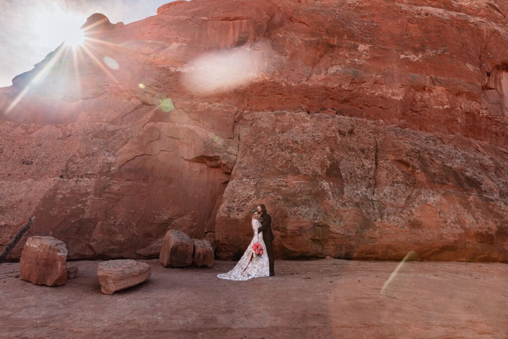 Couple holding each other in front of red rock at Look Glass Arch Moab elopement, with sunflare creating a warm golden glow over the desert landscape.