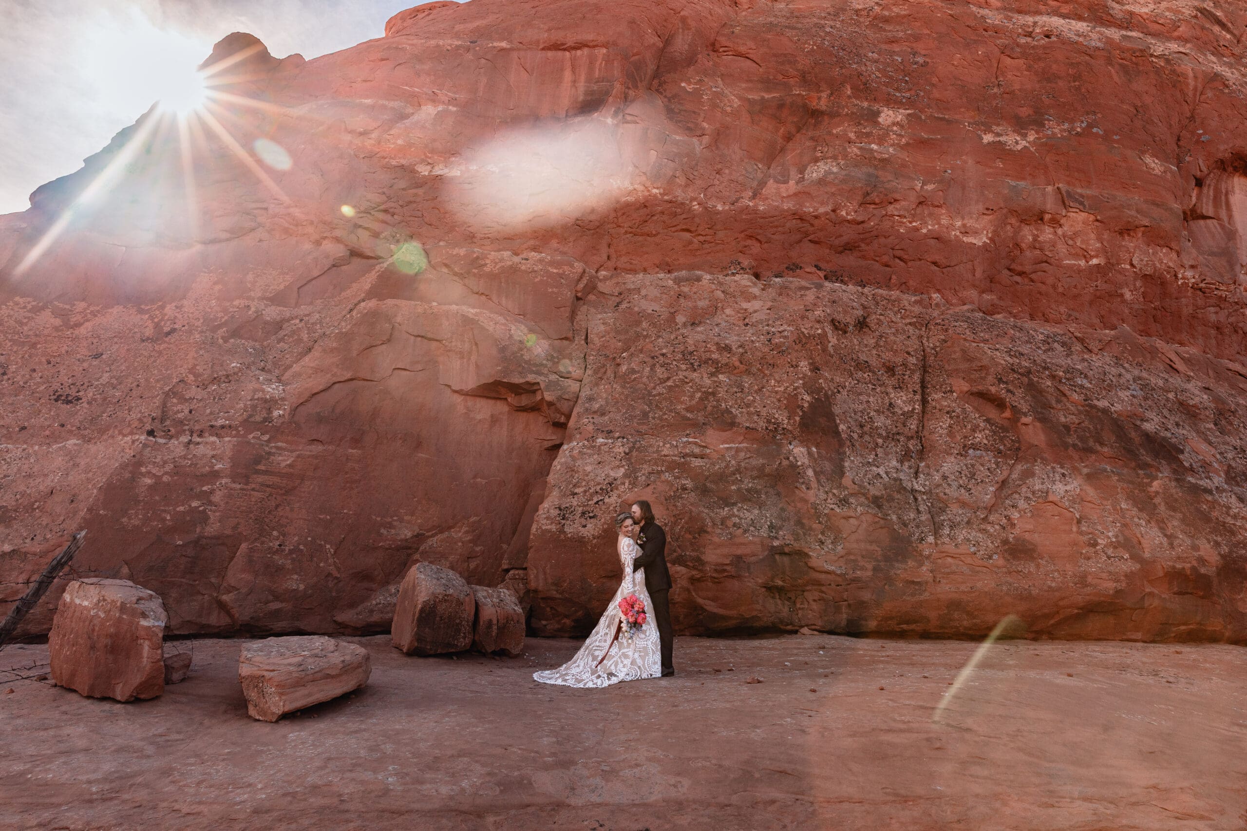 Couple holding each other in front of red rock at Look Glass Arch Moab elopement, with sunflare creating a warm golden glow over the desert landscape.