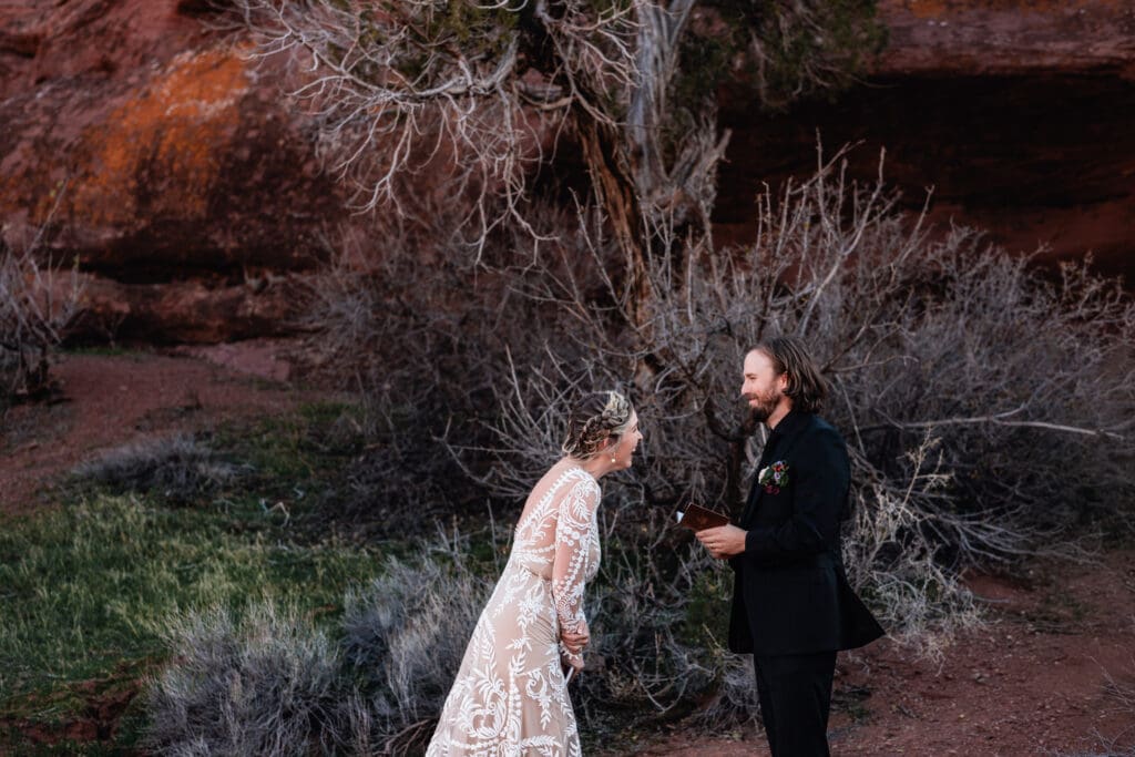 Bride laughing at her groom’s vows during a Moab elopement ceremony, surrounded by red rock cliffs and golden sunset light, capturing candid desert wedding joy.