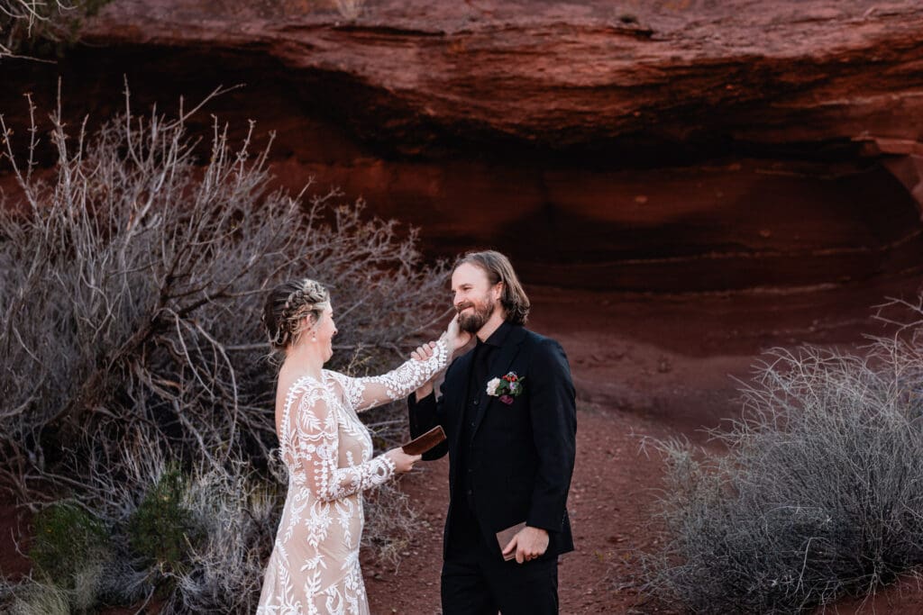 Bride tenderly caressing her groom’s face while sharing vows at a Moab wedding, with iconic red rock formations creating a romantic desert backdrop.