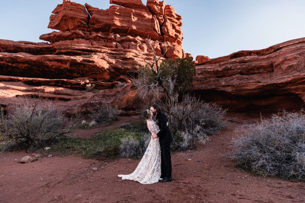 Couple sharing their first kiss as newlyweds at a Moab elopement, celebrating under a colorful desert sunset with red rock cliffs in the background.
