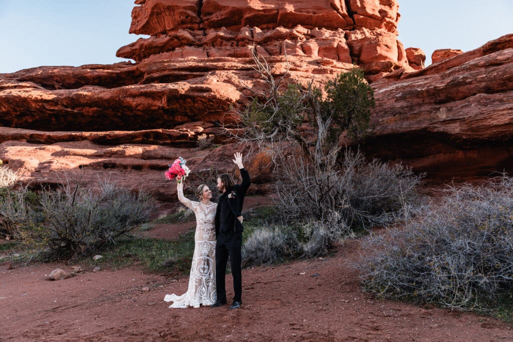 Newlywed couple celebrating after being pronounced husband and wife during a Moab elopement, surrounded by red rock cliffs and a golden desert sunset.