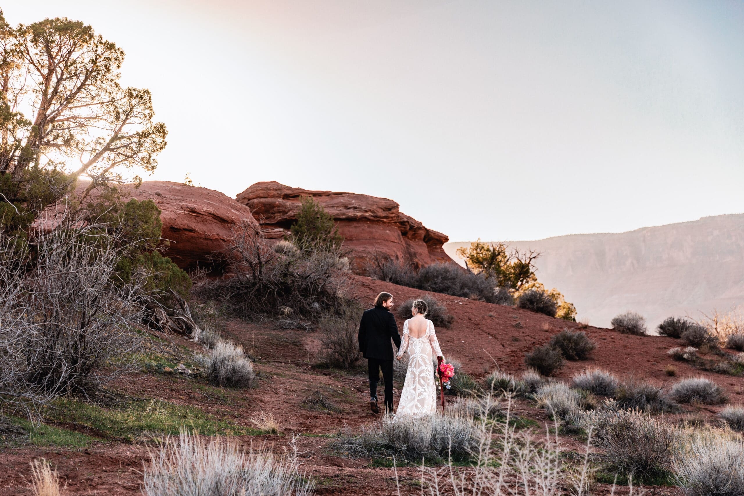 Newly married couple walking hand in hand across the red rock landscape after their intimate Moab elopement ceremony.