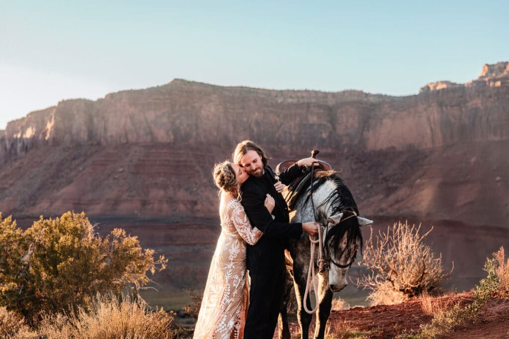 Bride hugging the groom from behind and kissing his cheek at sunset with a horse in the background during their Moab desert elopement.