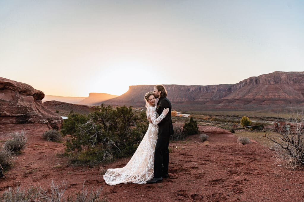 Groom kissing his bride’s forehead during a Moab horse ranch elopement at sunset, with golden light dipping behind red rock canyons and a romantic desert backdrop.