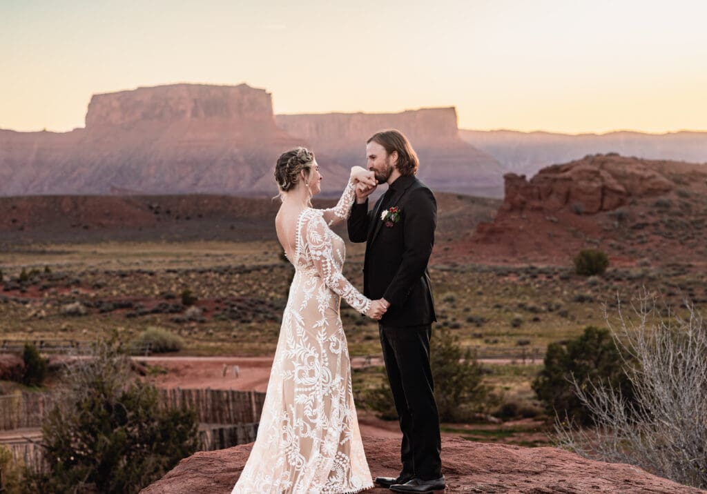 Groom kissing his bride’s hand during a Moab elopement at sunset, with canyons and red rock mesas creating a dramatic desert backdrop.
