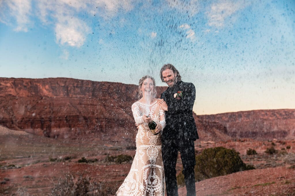 Newlywed couple popping champagne at a Moab horse ranch elopement at sunset, celebrating with red rock desert scenery and golden light in the background.