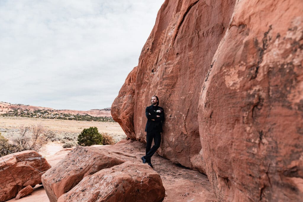 Groom in a black suit leaning against red rock at a Look Glass Arch Moab elopement, with sweeping desert views in the background.
