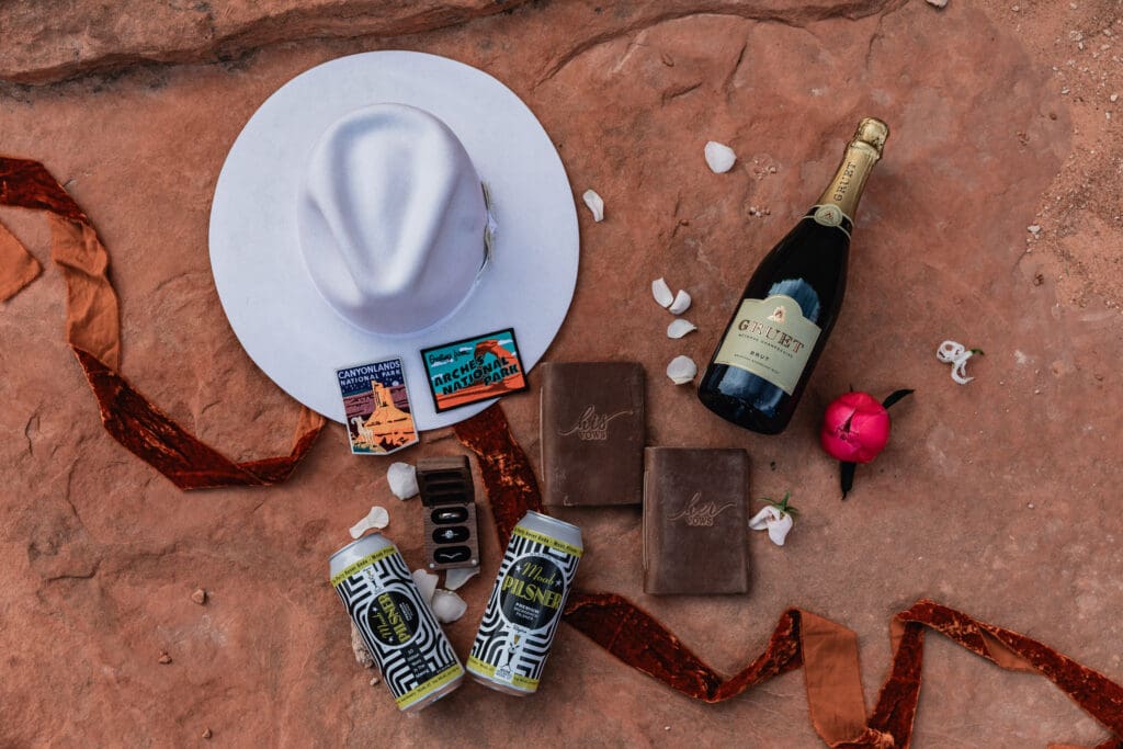 Flat lay of elopement details including Moab beer, white hat, vow books, and wedding rings arranged for a desert elopement detail photo.