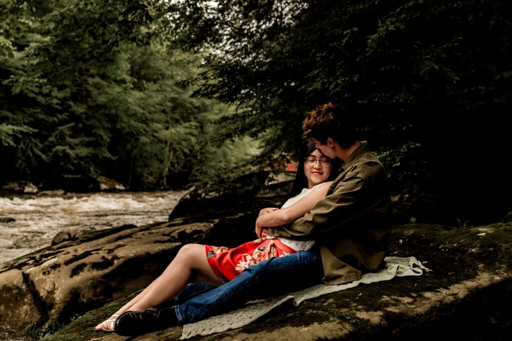 Couple sits and snuggles together by a flowing stream during a romantic engagement photo session at McConnells Mill State Park