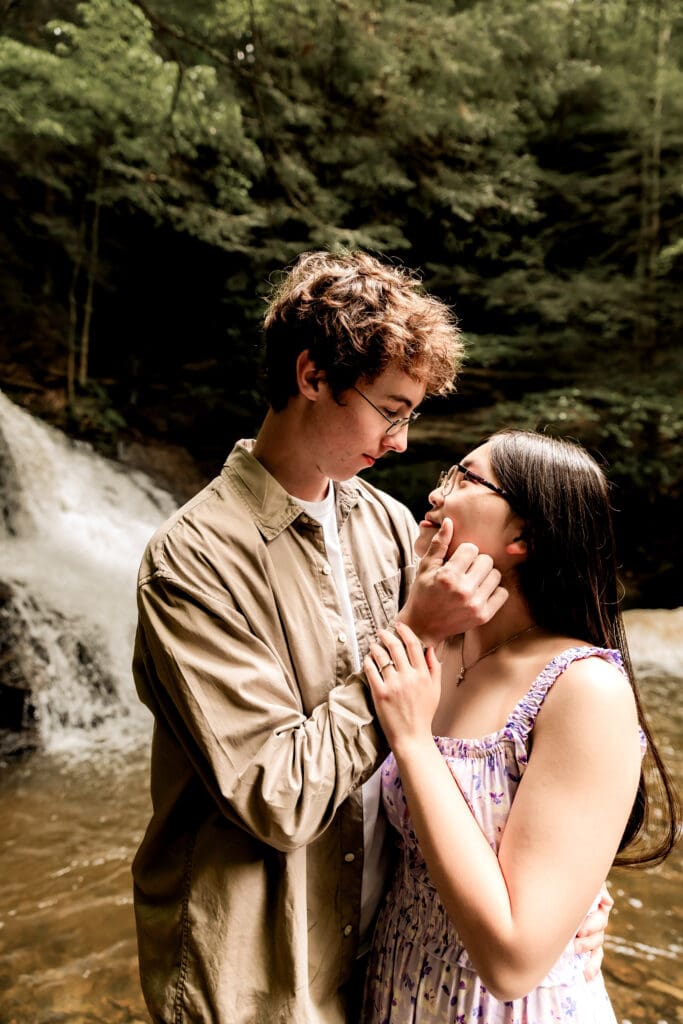 Couple embraces near a cascading waterfall in a lush forest during a romantic engagement photoshoot at McConnells Mill State Park
