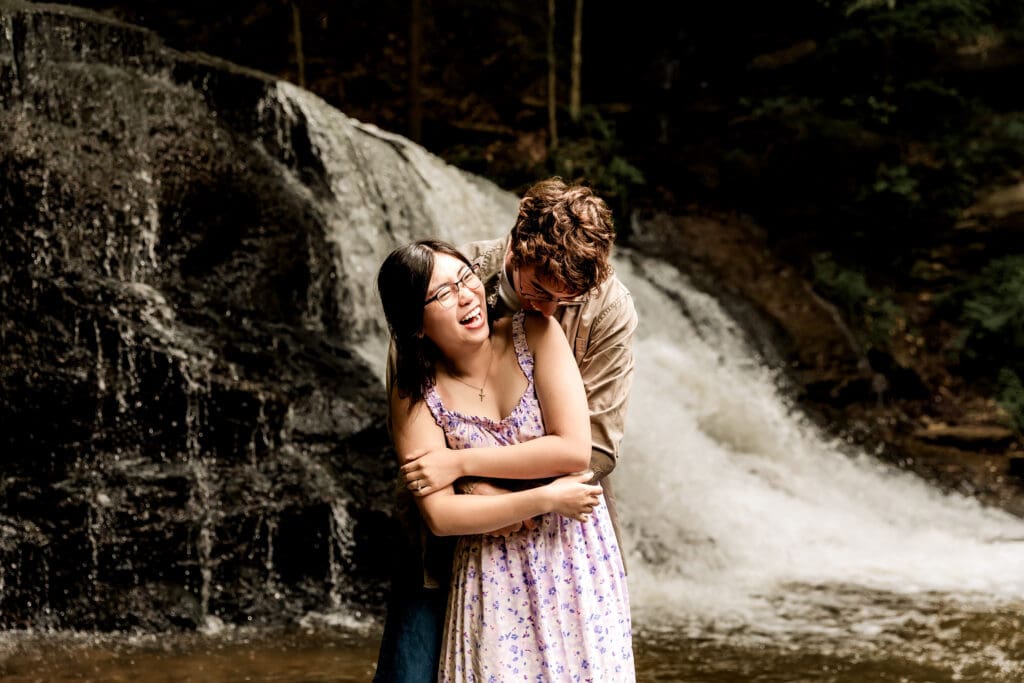 Romantic couple embraces by a forest waterfall at McConnells Mill State Park, wearing a beige jacket and purple floral dress