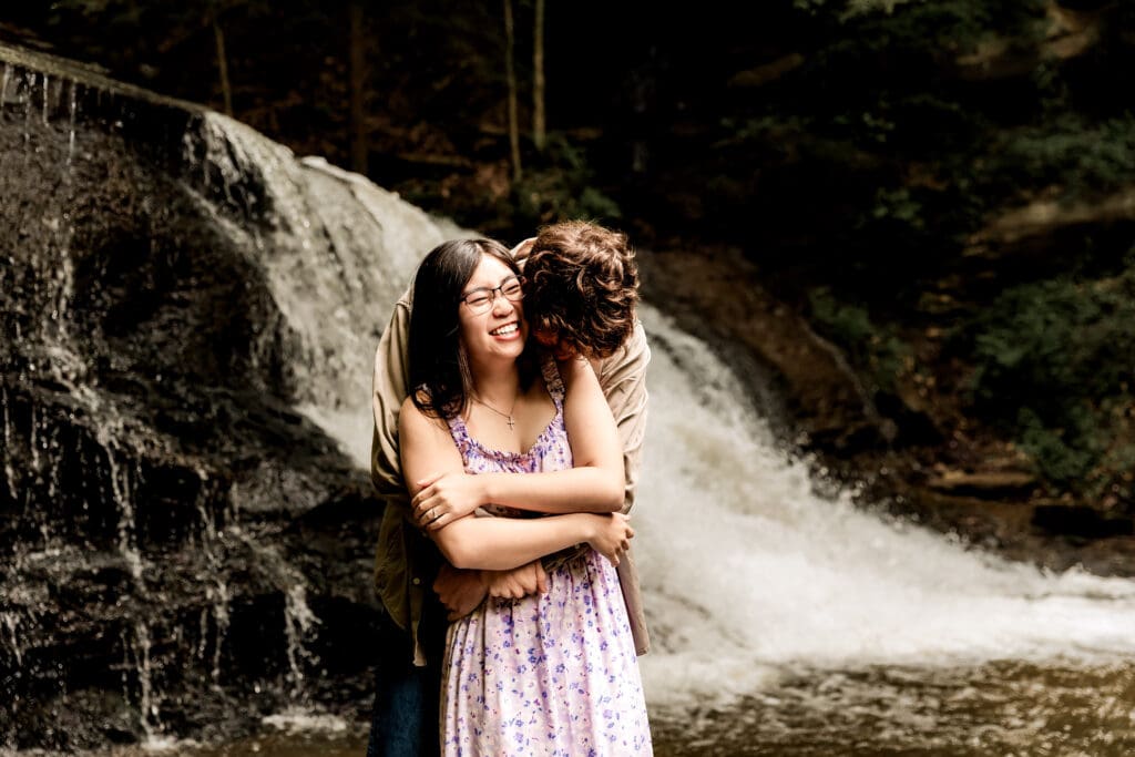 Couple embraces in front of a cascading forest waterfall at McConnells Mill State Park, wearing a floral dress and beige outfit