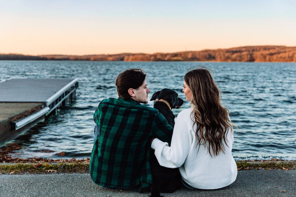 Two people sit together with their dog on a dock overlooking a serene lake during sunset engagement photos
