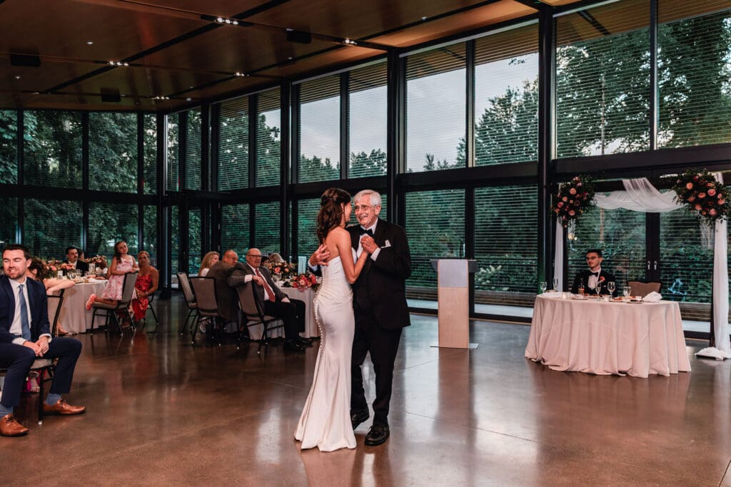 Bride dancing with her father in elegant glass-walled reception venue at the National Aviary