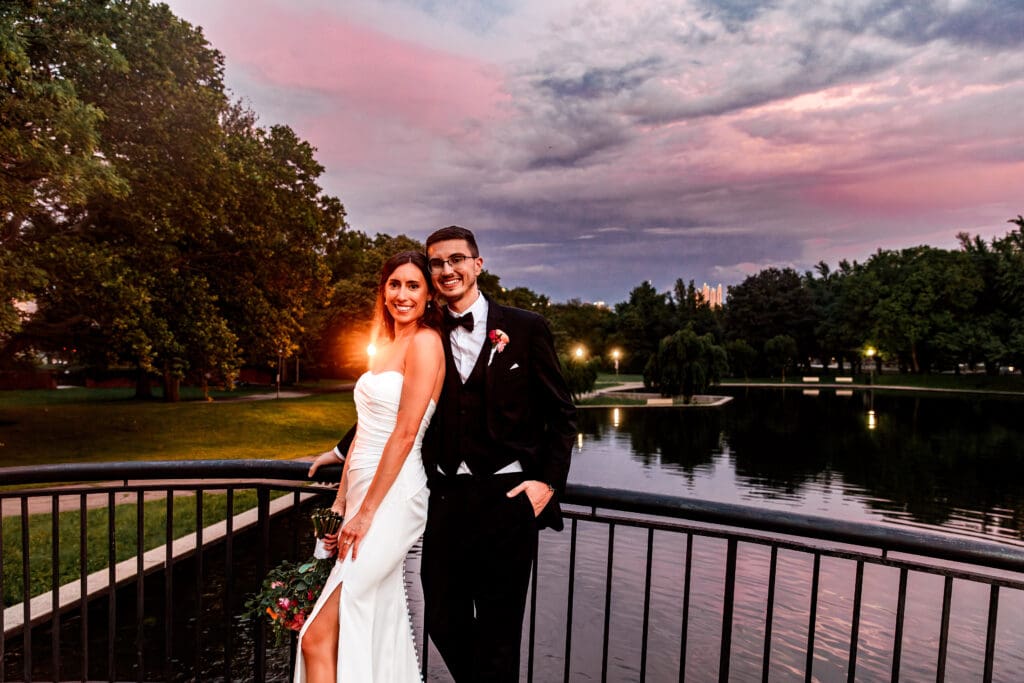 Wedding couple posing together at lakeside sunset with dramatic pink and purple clouds at the National Aviary